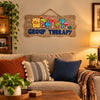 A decorative wooden wall sign with a wavy edge, featuring colorful cocktail glasses and the words 'GROUP THERAPY' in blue letters, hanging against a plain light gray wall.